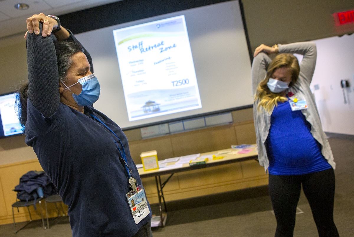 Two women wearing surgical masks and lanyards with nametags pull their elbows as they stretch their arms. A slide with the words “Staff Retreat Zone” is on a screen between them. A table is in the background.