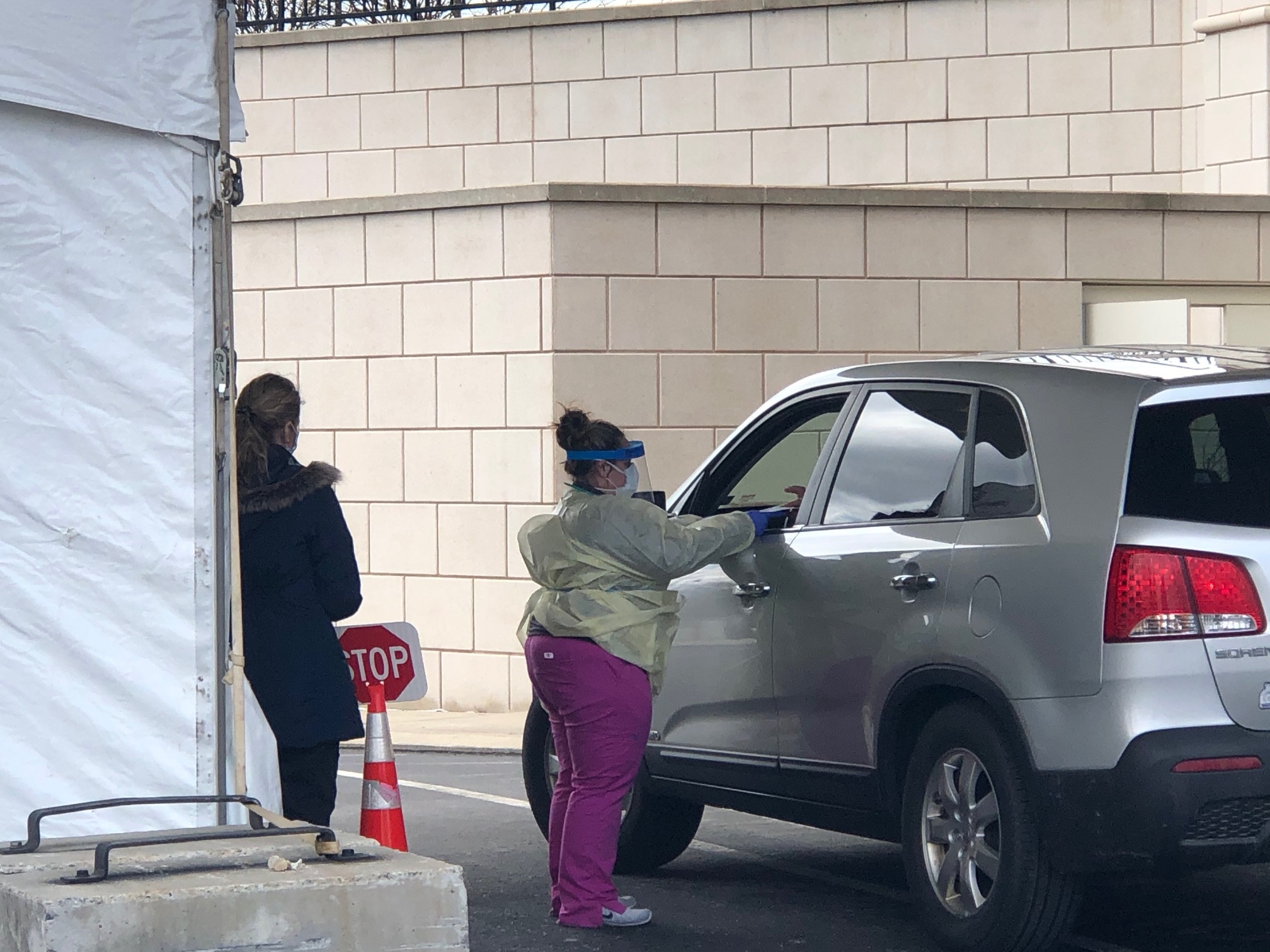 A woman dressed in scrubs with a face mask and shield hands a paper through a car window to a patient who has arrived for a COVID-19 test. Another woman stands behind her, learning on a tent.