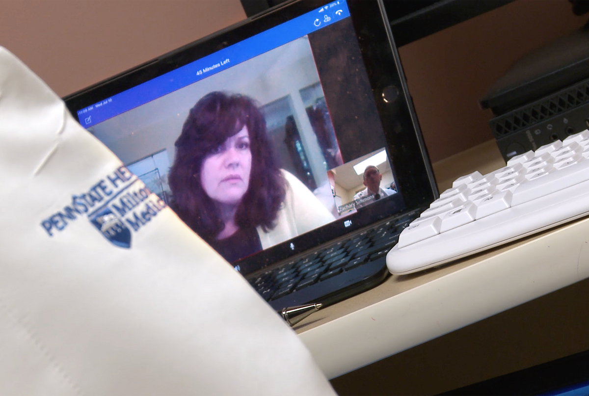 Cynthia Lacey is seen in a close-up on a laptop screen. She has medium-length hair and is wearing a sweater. Dr. Zachary Simmons’ sleeve is in the foreground.