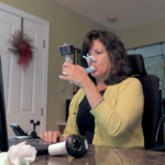 A woman holds a device called a spirometer to her mouth as she looks at a laptop screen. She has medium-length hair and is wearing a sweater. A man is standing to her right.