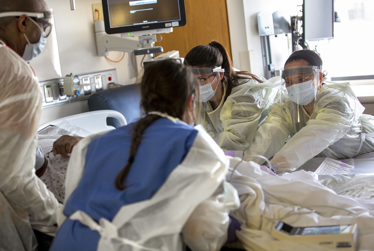 Nancy Rudy and Judylyn Silot, facing camera, from left, registered nurses with the Medical Intermediate Care Unit at Hershey Medical Center, and two other health care workers, whose backs are to the camera, touch a patient who is lying in bed. They are wearing gowns and face shields. Behind them is a monitor.