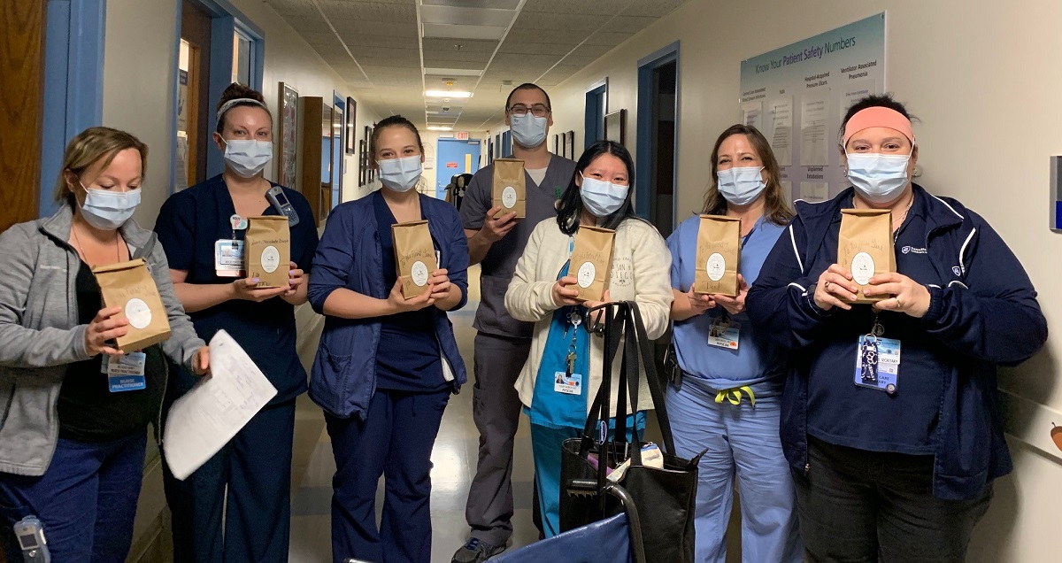 Seven members of the Neonatal Intensive Care Unit are holding bags of coffee. They are all wearing scrubs and masks and standing in the hallway of their unit.