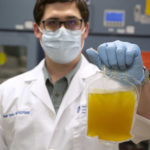 A man wearing a lab coat, gloves and face mask holds up a bag of plasma as he looks at the camera.
