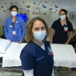Registered nurses Jamie Boyer, Carol Brown and Kristin Gardyasz, dressed in scrubs and wearing face masks, are standing beside a bed in an exam area. There are stickers and pictures of animals on the walls.
