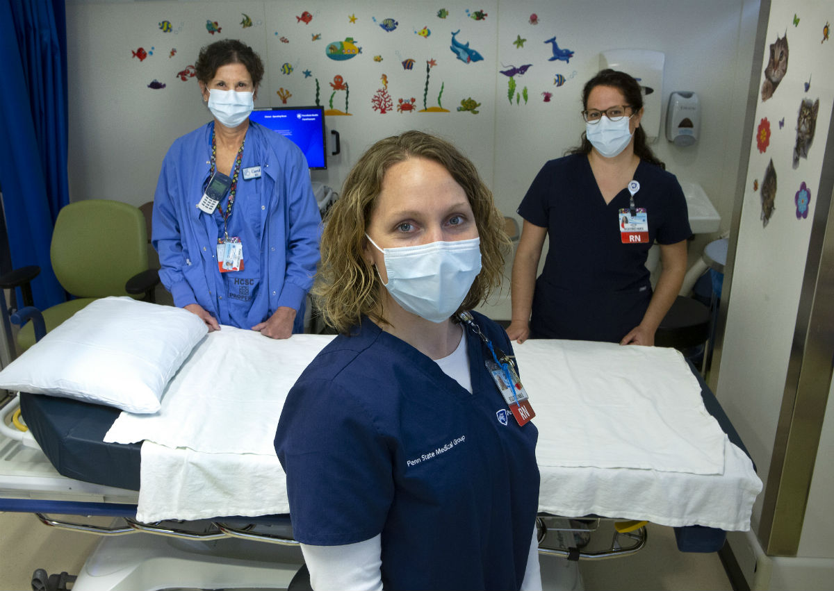 Registered nurses Jamie Boyer, Carol Brown and Kristin Gardyasz, dressed in scrubs and wearing face masks, are standing beside a bed in an exam area. There are stickers and pictures of animals on the walls.