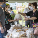 A woman wearing a mask takes a bag of items from another woman, also wearing a mask, at an outdoor farmers' market. Other people with masks look on.