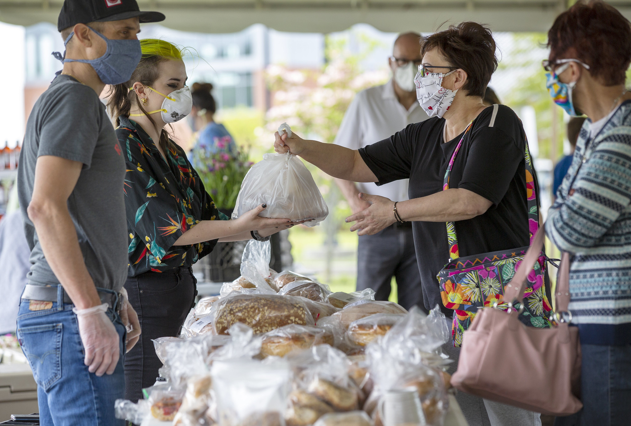 A woman wearing a mask takes a bag of items from another woman, also wearing a mask, at an outdoor farmers' market. Other people with masks look on.