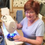Director of volunteer services Barbara Moyer sits at her sewing machine as she makes a face mask. She is wearing a purple short sleeve shirt, gray pants and glasses.