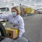 La enfermera registrada Stephanie McGaw, que lleva una sudadera, guantes y una máscara, levanta una caja de cartón de comida en la cajuela de un automóvil. Un camión con remolque y conos de tráfico que marcan los carriles de tránsito están en el estacionamiento detrás de ella.