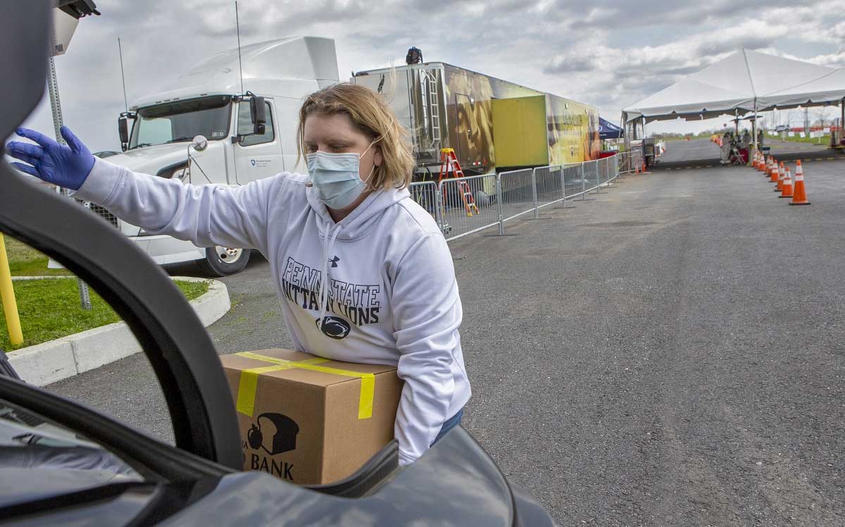 La enfermera registrada Stephanie McGaw, que lleva una sudadera, guantes y una máscara, levanta una caja de cartón de comida en la cajuela de un automóvil. Un camión con remolque y conos de tráfico que marcan los carriles de tránsito están en el estacionamiento detrás de ella.