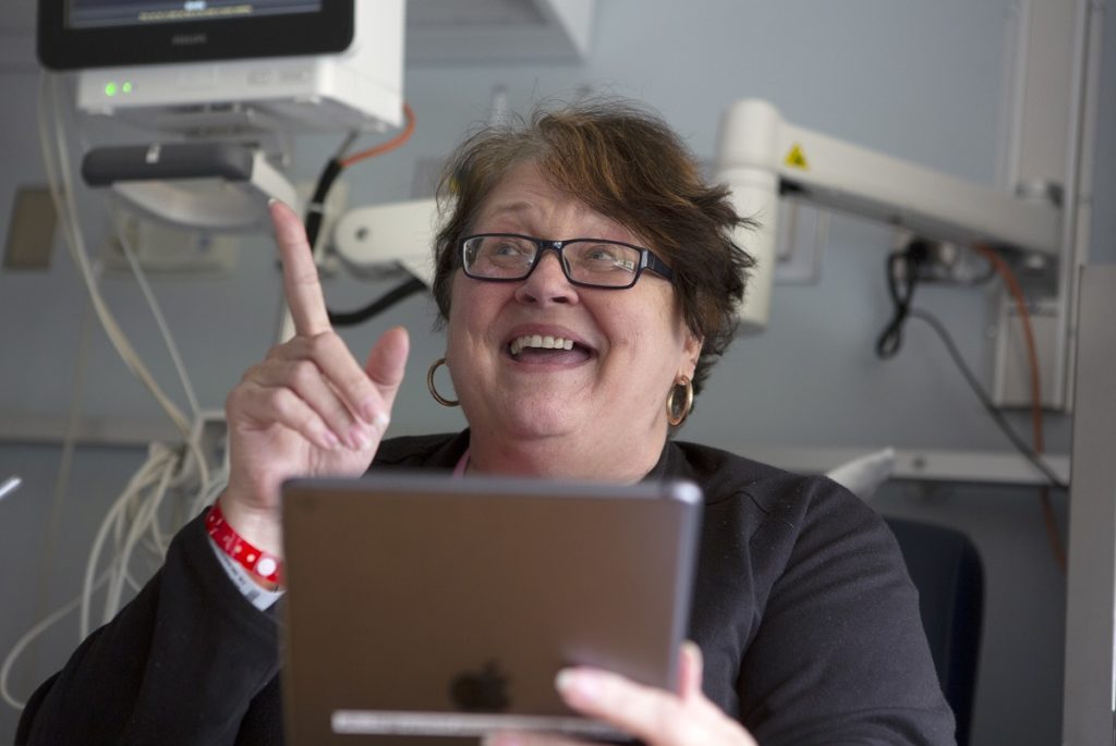 Patient Monica Chincharick points her index finger upwards as she smiles while holding an iPad. She has short hair and glasses and sits in front of hospital equipment in her room.