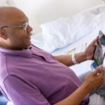 Patient Maurice Brown, who is bald and wears glasses, sits in a chair and smiles down at an iPad in his hands. The screen shows the smiling face of medical student Rahul Gupta. A hospital bed and window are in the background.