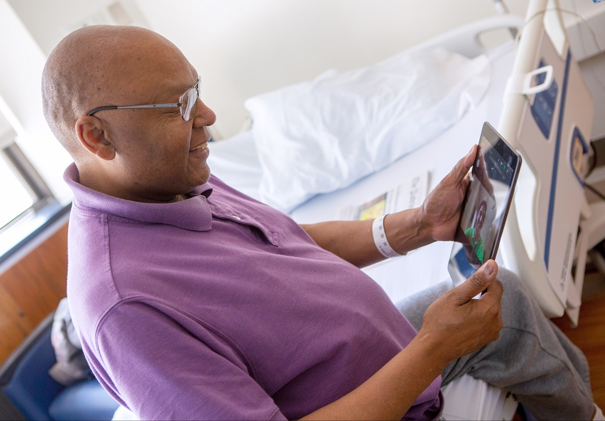 Patient Maurice Brown, who is bald and wears glasses, sits in a chair and smiles down at an iPad in his hands. The screen shows the smiling face of medical student Rahul Gupta. A hospital bed and window are in the background.