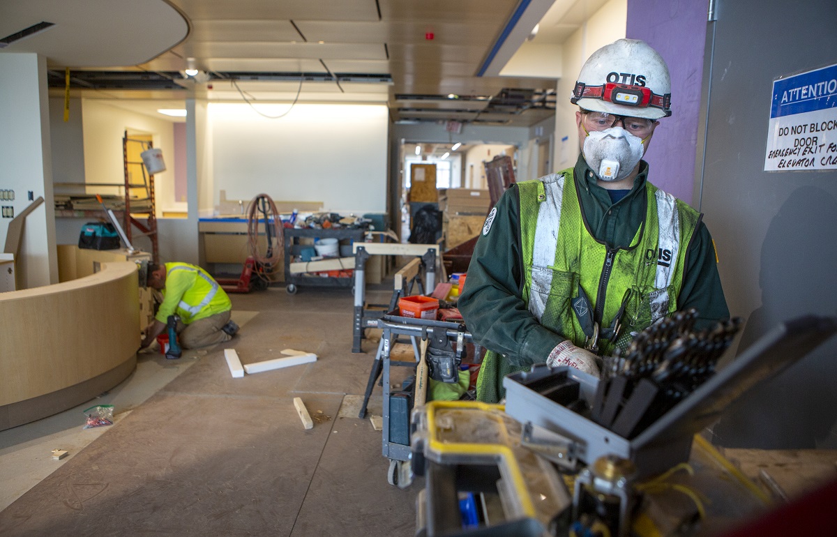 Two construction workers work on the Penn State Health Children's Hospital expansion in April. The man on the right is wearing a safety helmet, N95 mask and safety vest. The man in the back is kneeling and working on the counter. Tools and equipment are scattered around.