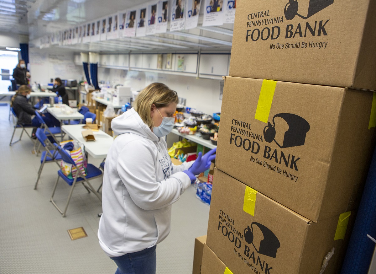 A woman wearing a mask, gloves and sweatshirt stands next to a stack of cardboard boxes from the Central Pennsylvania Food Bank. Behind her is a row of tables in a warehouse and three people out of focus.