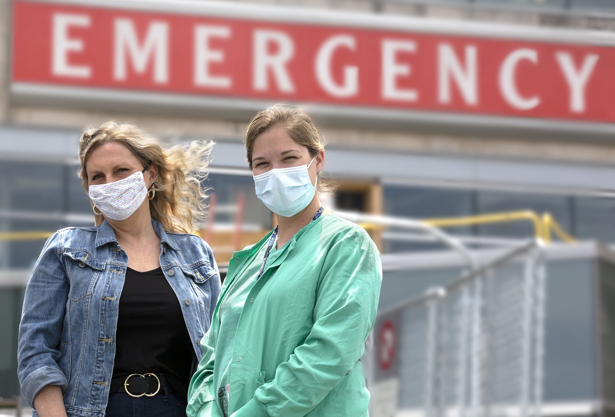 Marie Conley and Kaitlin Snook stand outside the emergency department entrance at Hershey Medical Center. Marie wears a jean jacket and has wavy hair. Kaitlin wears scrubs with her hair in a ponytail. Both wear masks. The word “emergency” is on a sign on the building.