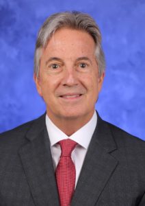 Frank smiles at the camera. He is wearing a suit and tie.