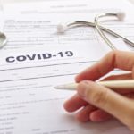 A hand holds a pen and stethoscope above a health insurance form with the word "COVID-19" at the top.
