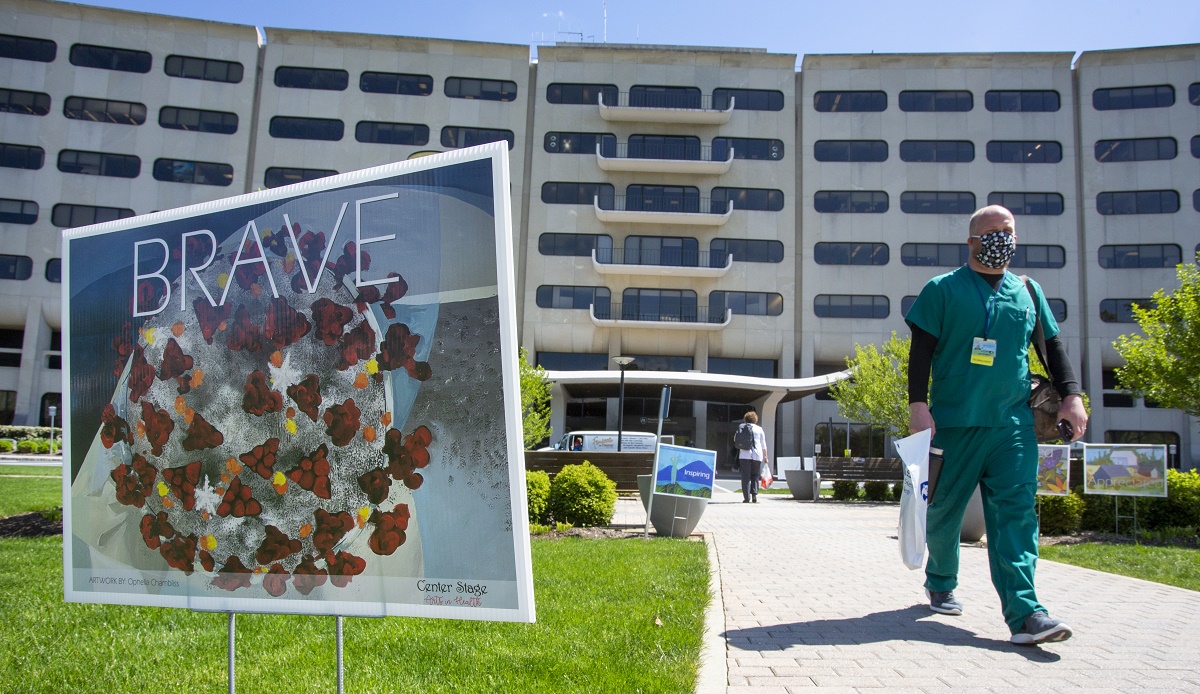 A health care worker dressed in scrubs and wearing a face mask walks past a curbside sign that shows a graphic of COVID-19 and says “Brave.” Hershey Medical Center is in the background.