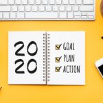 A day planner book with “2020” and the words “Goal, plan, action” sits on a desk surrounded by a keyboard, plant, post-it notes, coffee, pens glasses and cell phone.