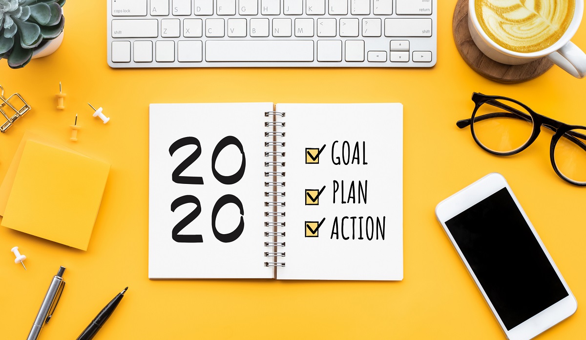 A day planner book with “2020” and the words “Goal, plan, action” sits on a desk surrounded by a keyboard, plant, post-it notes, coffee, pens glasses and cell phone.