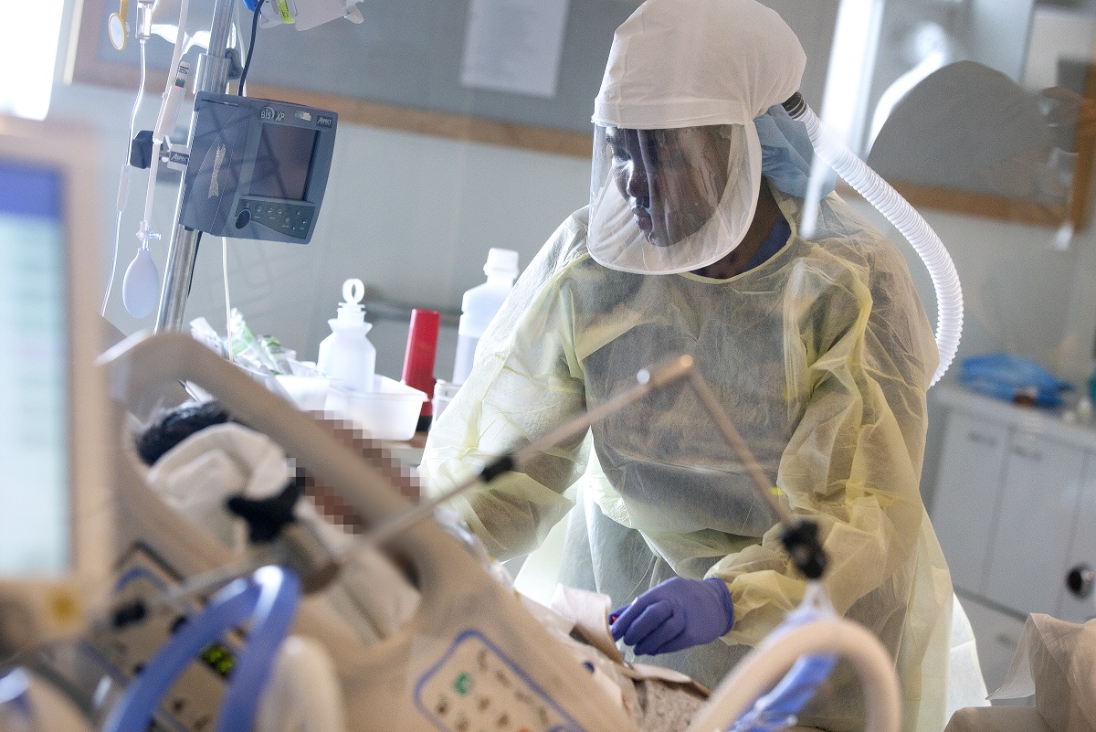 Mary Kamau, dressed in a gown, gloves and face mask, leans over the bed of a patient whose face cannot be seen. Medical equipment mounted on an IV pole can be seen in the background.