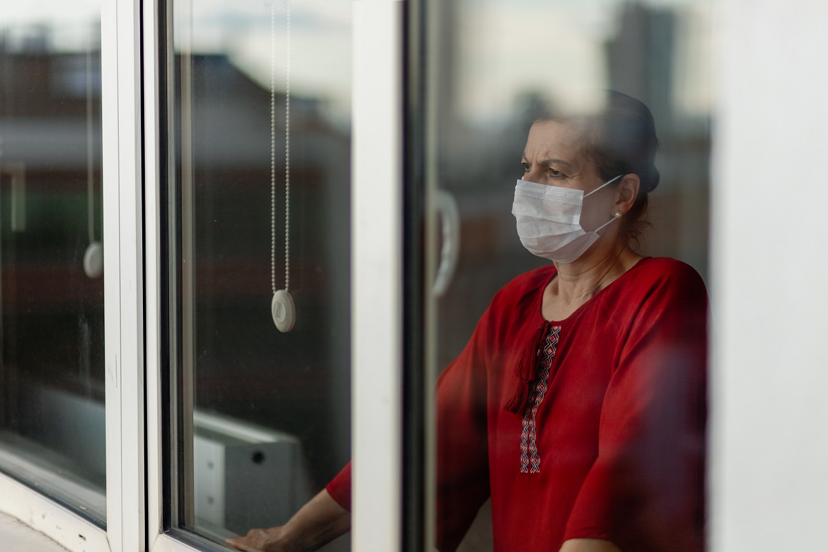 A woman wearing a mask looks out a window. Her brows are furrowed and she looks worried.]