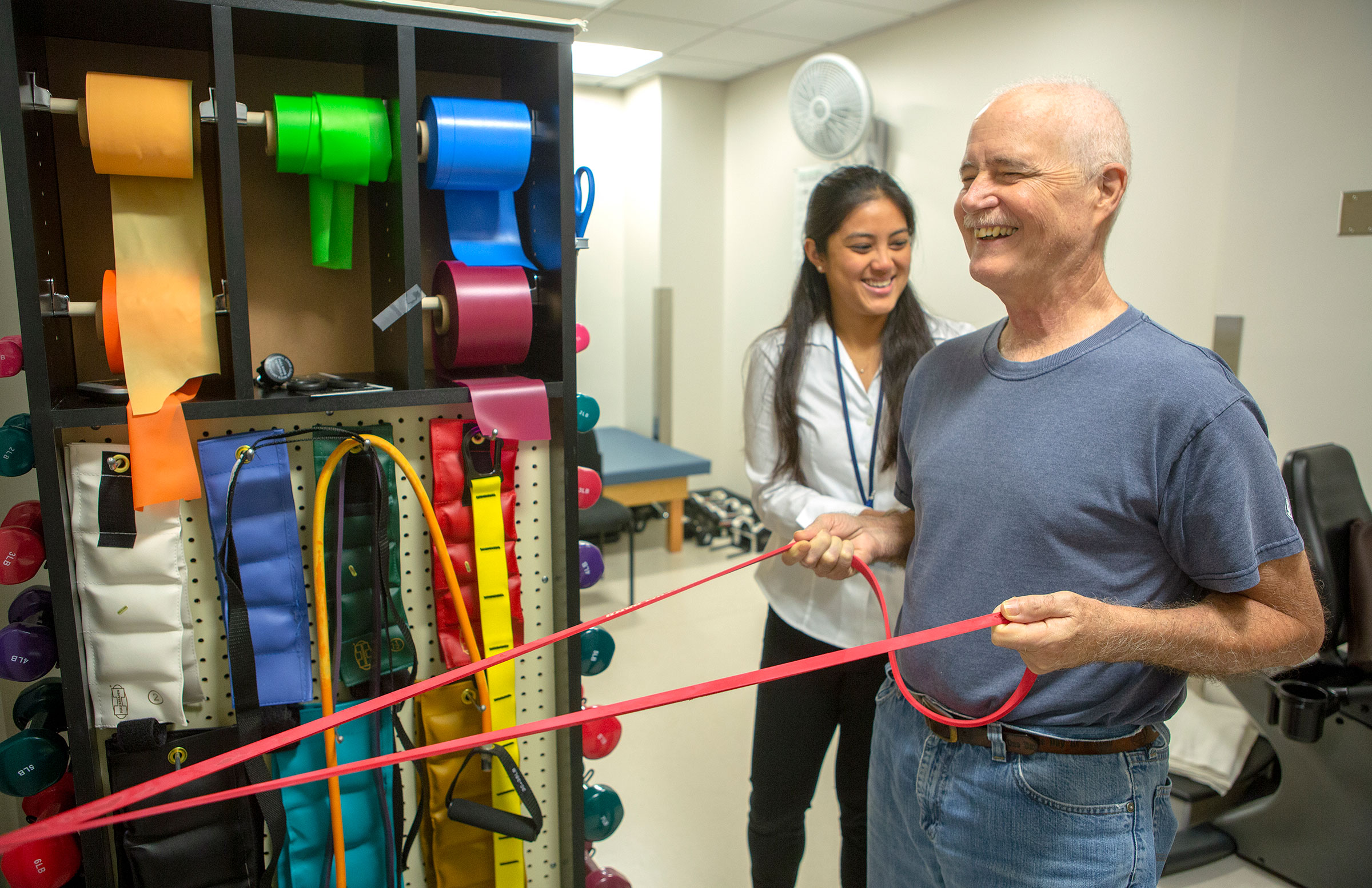 Melanie Potiaumpai, PhD, a postdoctoral scholar with The ONE Group, works with cancer patient Curt Chambers in the exercise room at Penn State Cancer Institute in 2018. Both are smiling and Chambers is pulling on a resistance band.