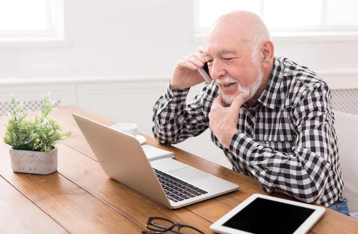 Senior man talking on phone and using laptop, sitting at table at home.