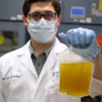 Un hombre con bata de laboratorio, guantes y mascarilla sostiene una bolsa de plasma mientras mira a la cámara.