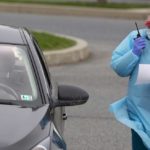 144/5000 Una enfermera que usa una bata, guantes y un protector facial sostiene un walkie-talkie mientras está parada junto a una mujer en un automóvil. La mujer lleva una máscara facial.