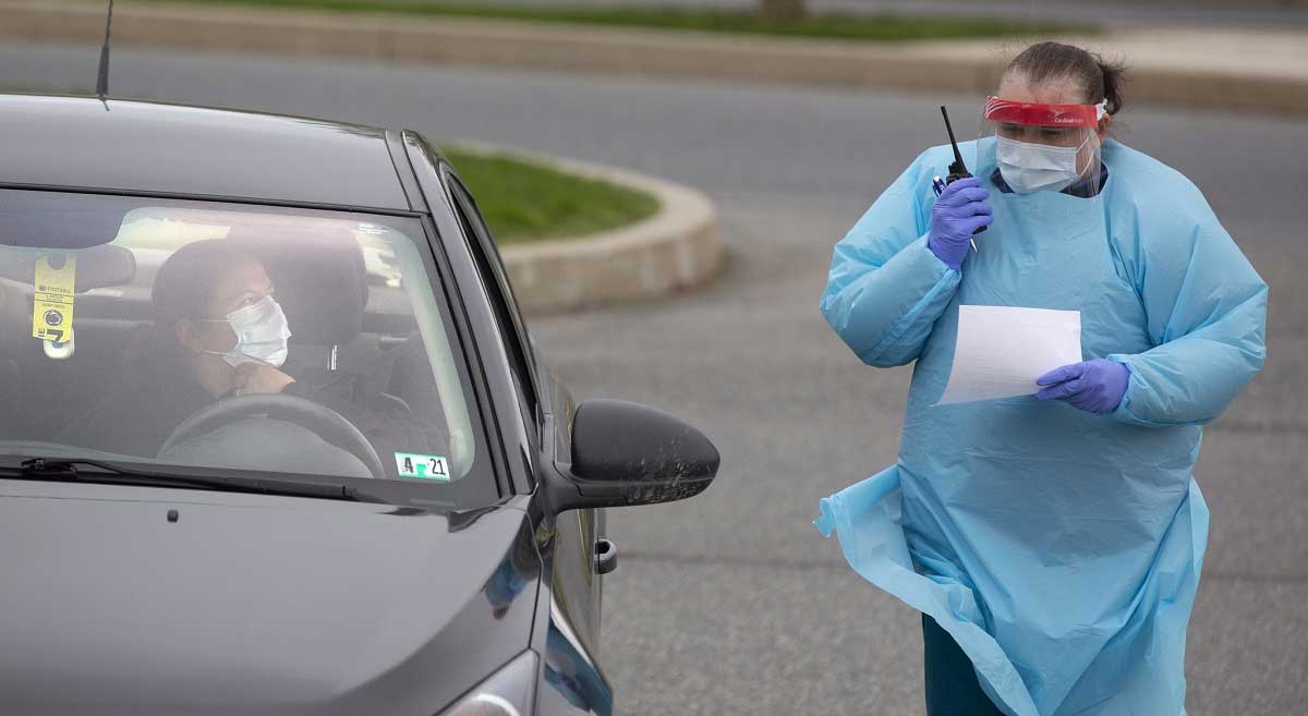 144/5000 Una enfermera que usa una bata, guantes y un protector facial sostiene un walkie-talkie mientras está parada junto a una mujer en un automóvil. La mujer lleva una máscara facial.