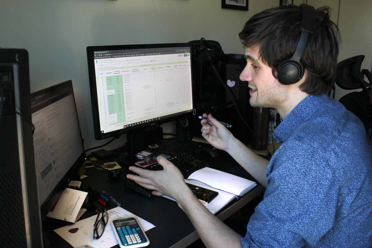Un hombre se sienta en un escritorio frente a dos monitores de computadora. Lleva auriculares y sostiene un bolígrafo en la mano derecha. Una calculadora, anteojos y otros artículos están en el escritorio.