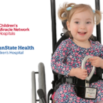 Miracle Child Stella holds a CMN fundraising award in her lap, as she sits in a wheelchair. The CMN Hershey and Penn State Health Children's Hospital logos appear to her left.