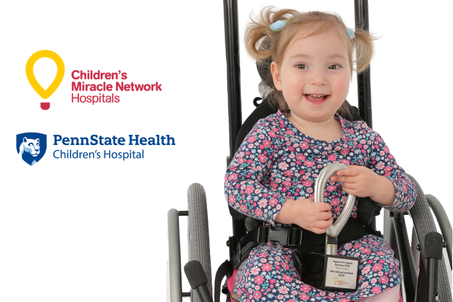 Miracle Child Stella holds a CMN fundraising award in her lap, as she sits in a wheelchair. The CMN Hershey and Penn State Health Children's Hospital logos appear to her left.