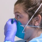 A nurse is seen in profile pinching the nose of an N-95 mask on her face. She is wearing rubber gloves, a smock and her hair is in a bun.