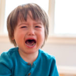 A young boy cries with his mouth open. In the background are some windows and furniture, slightly out of focus.