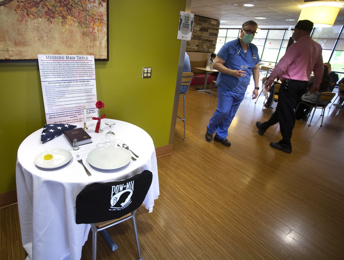 A fully set table is seen in the middle of a cafeteria. The chair is empty and has a POW-MIA banner. A folded American flag, Bible, single rose, candle and sign explaining the project are visible.