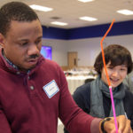 A student attempts to connect straws with small sticky blobs while two other students look on.