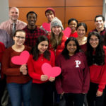A photo shows a group of 15 people wearing various shades of red and holding cut-out paper hearts.