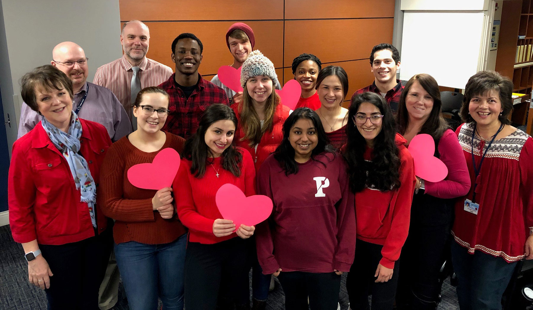 A photo shows a group of 15 people wearing various shades of red and holding cut-out paper hearts.