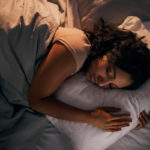 A woman lays in bed on her left side, under a comforter. Her eyes are closed and her arms are wrapped around a pillow.