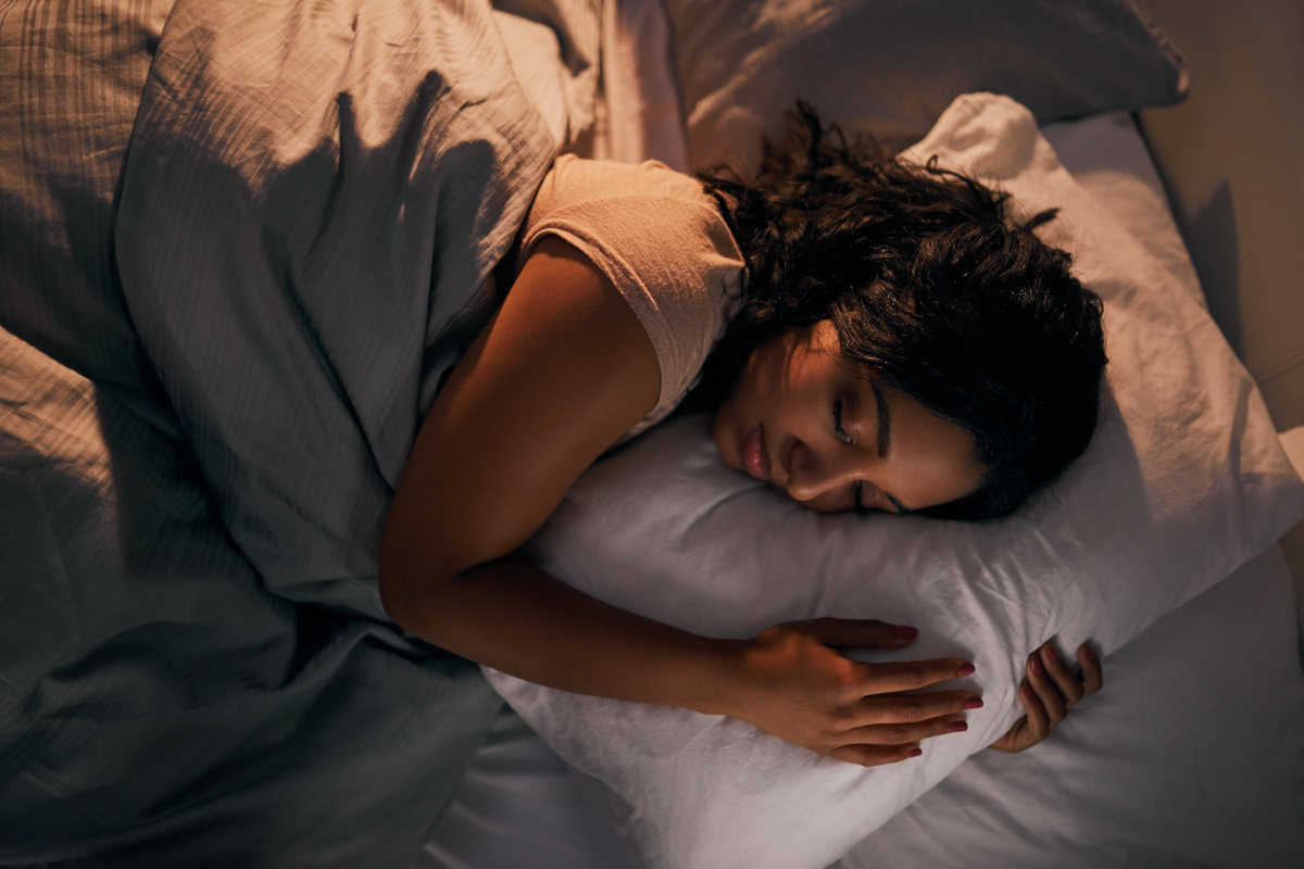 A woman lays in bed on her left side, under a comforter. Her eyes are closed and her arms are wrapped around a pillow.