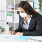 A woman wearing a mask sits at her desk. She’s holding a pencil in one hand and using a calculator with the other. Books, papers, a mug and bottle of sanitizer sit on top of the desk.