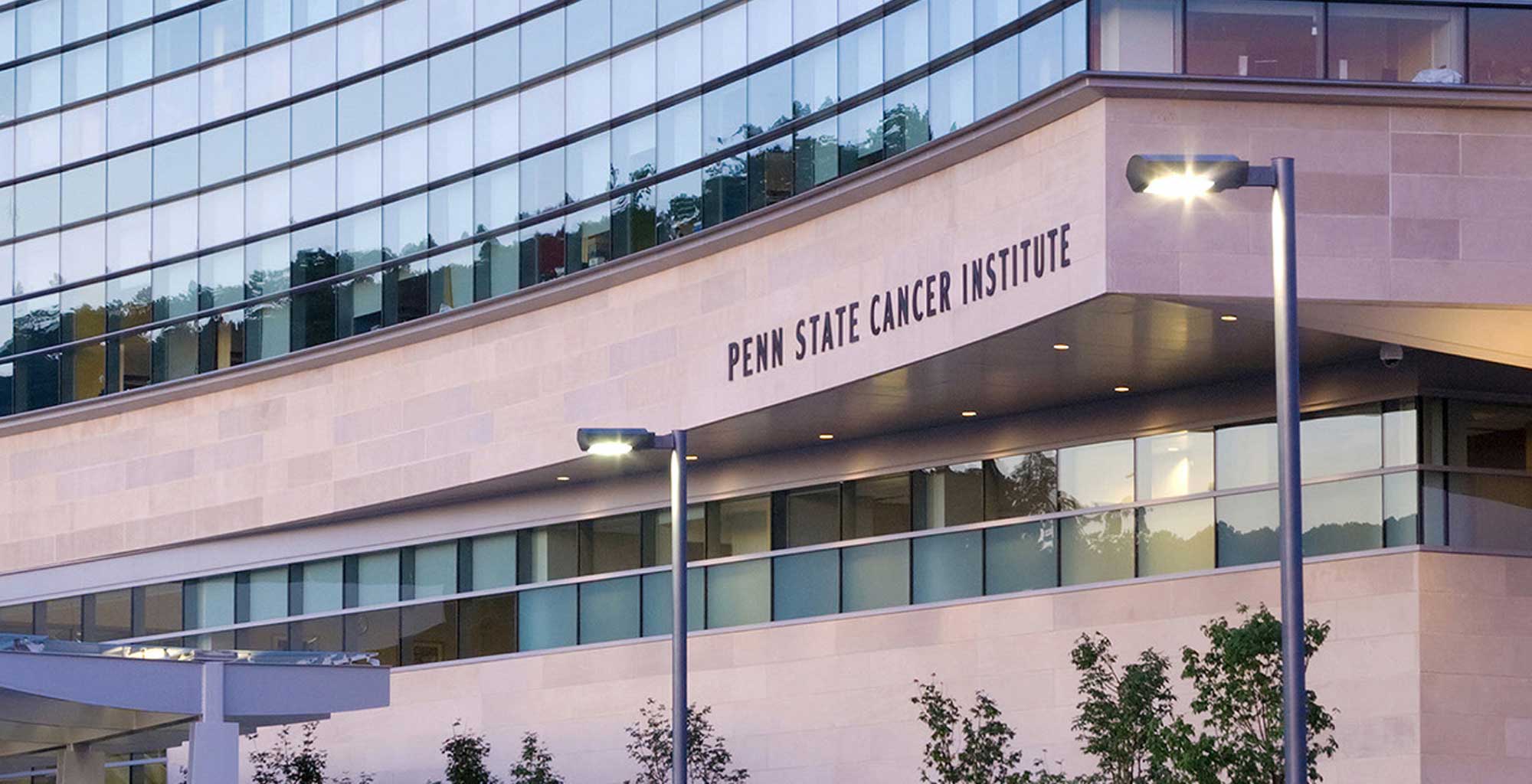 The Penn State Cancer Institute building is seen in the evening. A road leads up to it and a wall of glass windows is visible above the entrance.