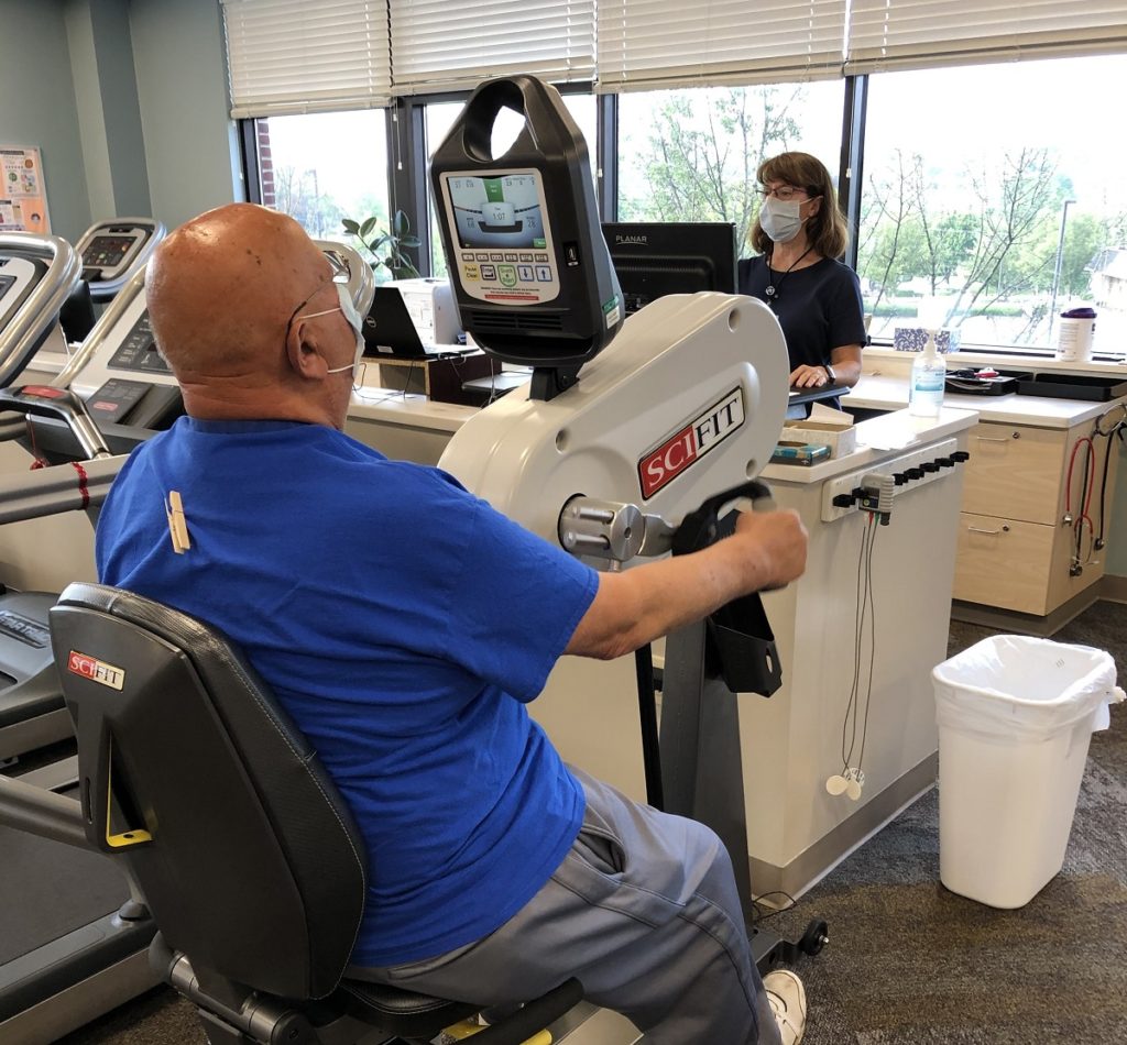A man sits at an arm bike with his arms in the pedals. A woman exercise physiologist look at a monitor as she connects him to a heart monitor. Both are wearing face masks.