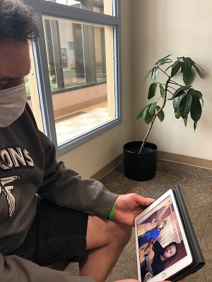 A person is seen wearing a face mask and holding an iPad. There is a video playing on the iPad.