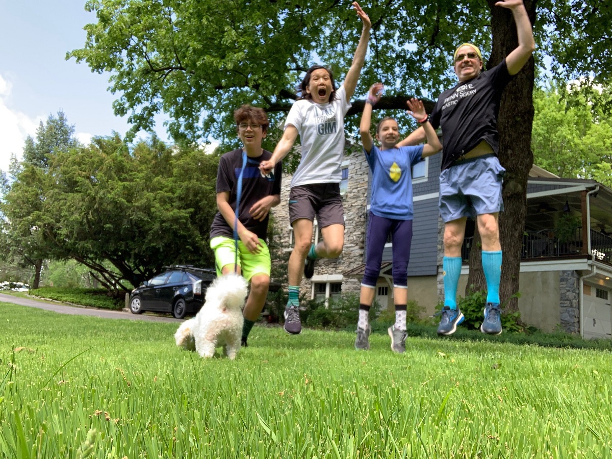 Four people jump off the grass outside of a house. One holds a dog on a leash.