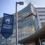 A blue and white flag hangs from a light pole in front of the entrance to Hershey Medical Center. On the flag is the Nittany Lion shield and the words, Penn State Health.]