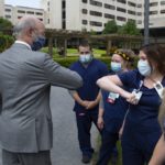 Gov. Tom Wolf, wearing a surgical mask and a suit, holds out an elbow to a woman in scrubs and a surgical mask, who offers her elbow in return. Three others in scrubs and surgical masks stand in a row. The widows and cement of Hershey Medical Center rise in the background.